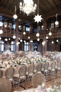 A grand hall filled with round-back wooden chairs with upholstered seats, arranged at elegantly decorated wedding reception tables with floral centerpieces and candles